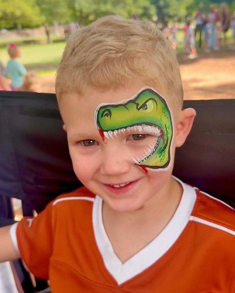 Smiling child with a detailed green dinosaur face painting by CandyProArt